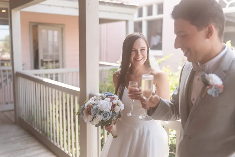 Sarah Katherine and Nathan toast with champagne on a sunlit veranda, holding a bouquet of white and blush flowers.