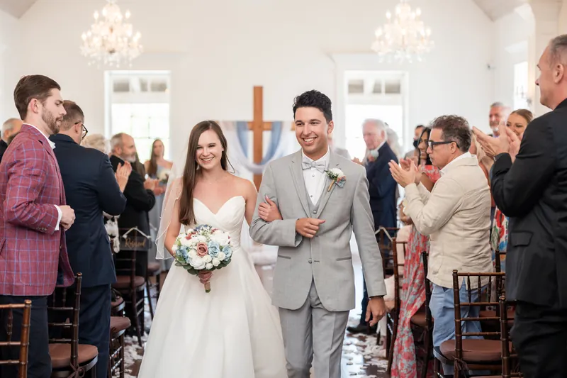 Sarah Katherine and Nathan walk back down the aisle together as guests applaud in the white-walled chapel.