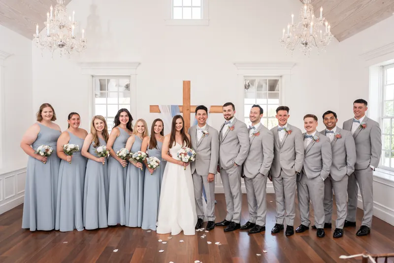 The wedding party stands in a single line in the bright chapel: attendants in slate blue dresses and groomsmen in gray suits flanking the couple.