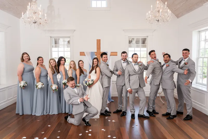 The full wedding party poses together in the chapel, with the couple at center and one groomsman crouched in front with a playful grin.