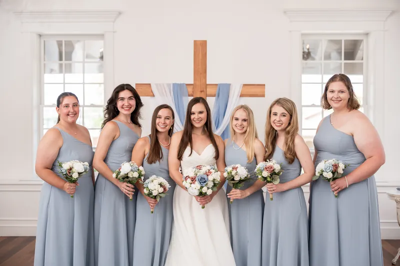 A person in a white gown stands with seven bridesmaids in slate-blue dresses, all holding white and blush bouquets before a wooden cross.