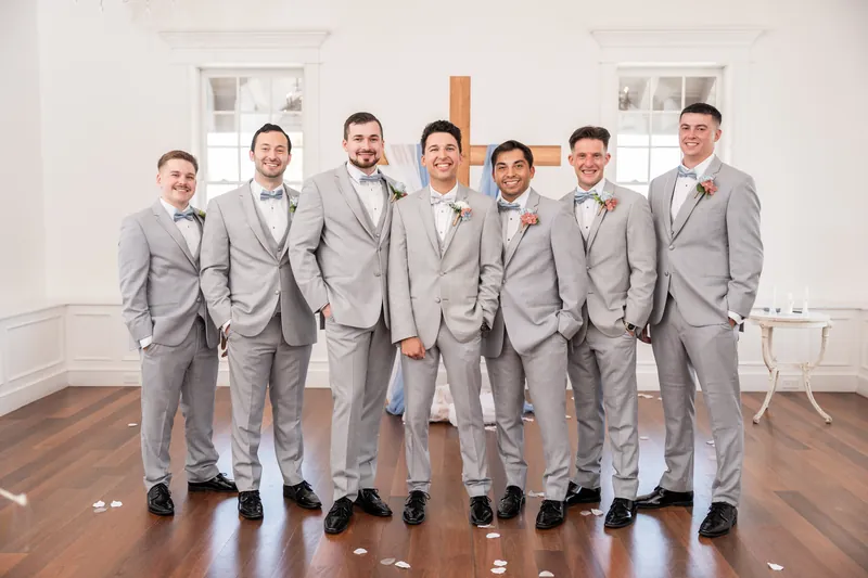 Seven groomsmen in matching gray suits stand shoulder-to-shoulder with hands in pockets in the chapel.