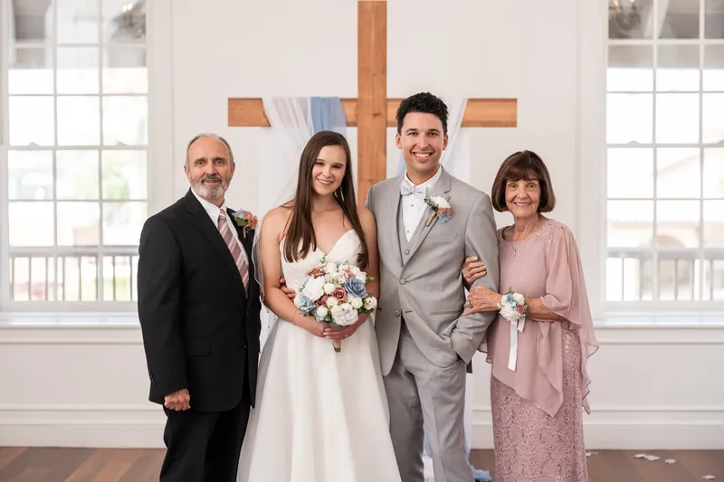 Four people pose together before the wooden cross, with two in wedding attire flanked by two in formal guest wear.