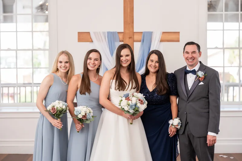 Four women in bridesmaid dresses and one man in a gray suit hold white bouquets beside the altar cross.