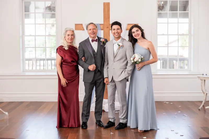 An older woman in maroon, two men in gray suits, and a person in a light blue dress stand together before the wooden cross.