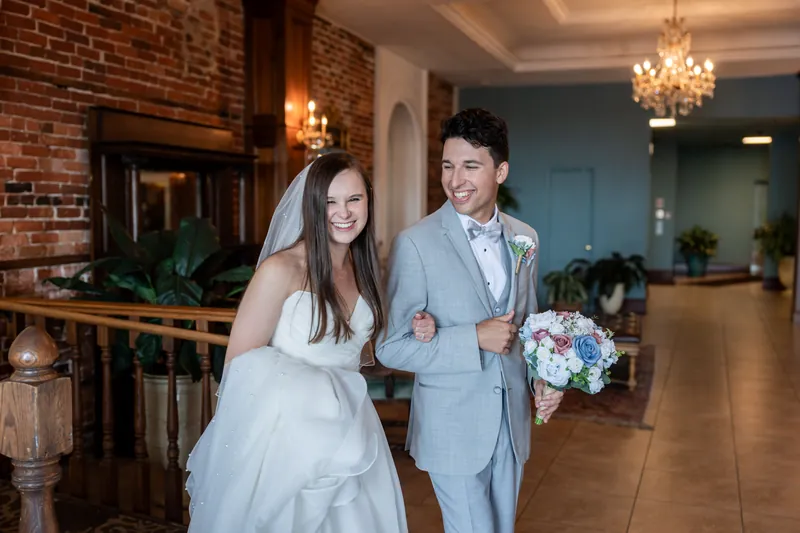 Sarah Katherine and Nathan walk arm-in-arm down the aisle at The White Room, she in a white gown with veil, he in a light blue suit, both smiling.