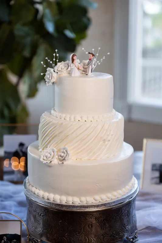 Three-tiered wedding cake with white frosting, diagonal piping on the middle tier, and a floral topper near a bright window.