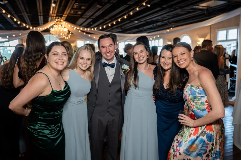 A group of six people in formal attire stand together at The White Room, with string lights and a chandelier overhead.