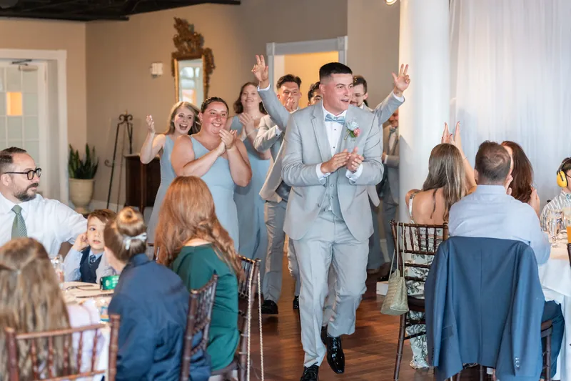 Two people in light blue suits walk down the aisle as guests cheer and raise their arms at The White Room.