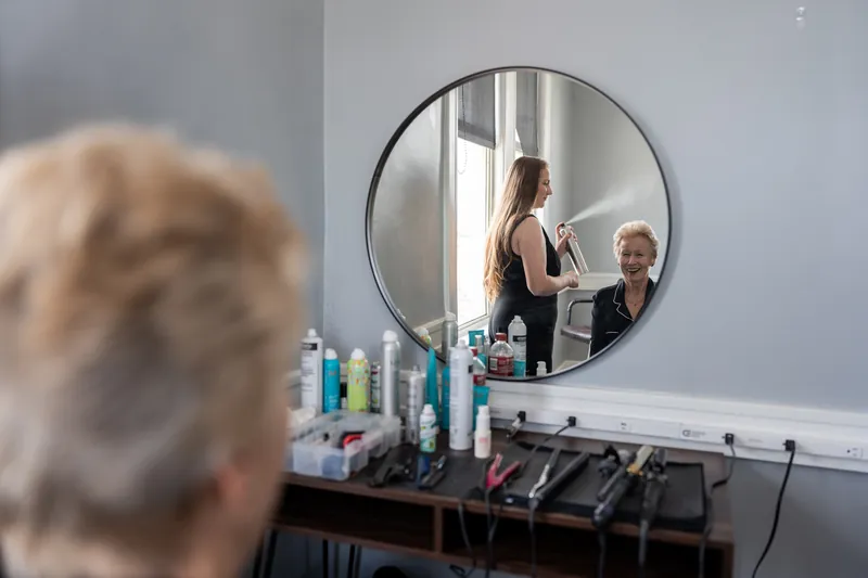 A woman in a white robe labeled "Mrs. Boone" adjusts her hair in a round mirror while cosmetics sit on the vanity below.