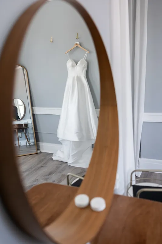 A white wedding dress hangs from a rod, reflected in an oval wooden mirror with candles on its surface.