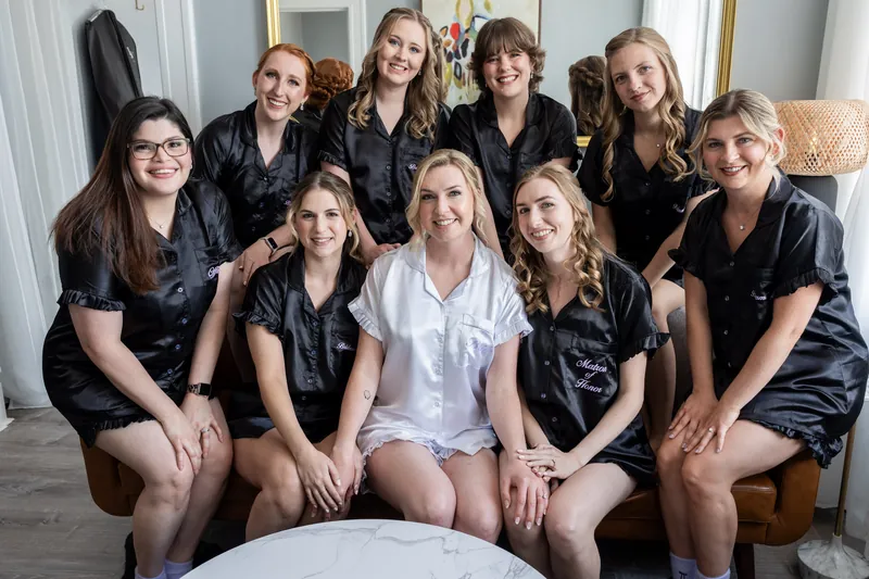 Sarah sits centered in white among ten women in matching black silk robes, all smiling at the camera in the bridal suite.