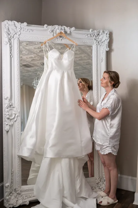 Two women adjust a white wedding dress hanging in an ornate mirror at The White Room.