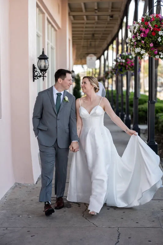 Sarah and Tim hold hands while walking along a columned portico adorned with hanging floral baskets at The White Room.
