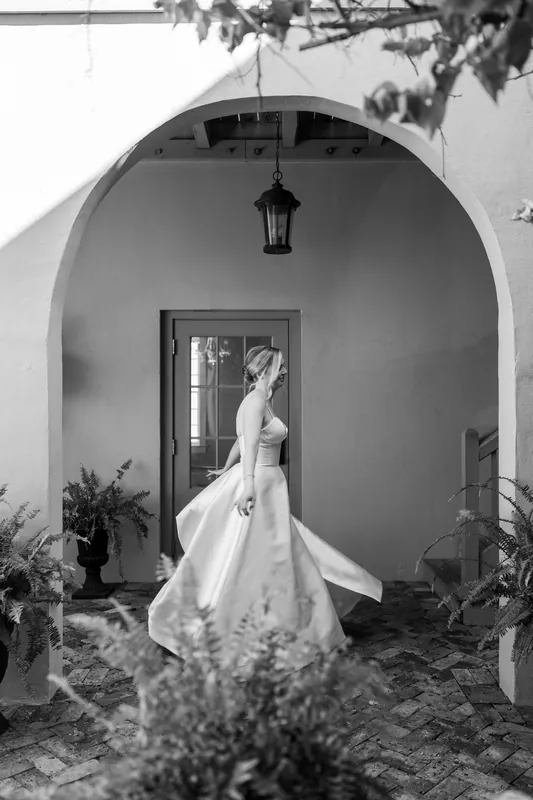 Sarah stands alone in a white gown, her dress train flowing, within a curved archway framed by potted plants.