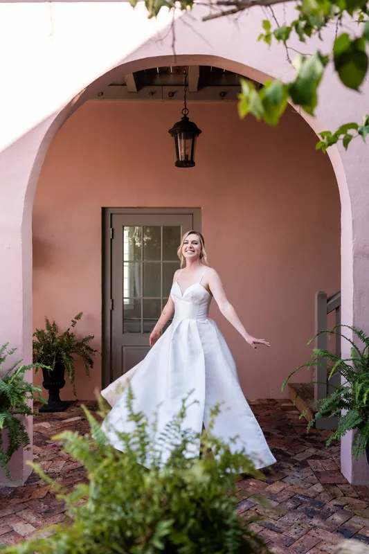 Sarah twirls in her white wedding dress beneath a pink arched entryway, foliage visible in the foreground.