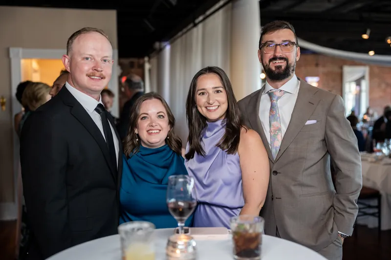 Two couples stand at a cocktail table during the reception, smiling at the camera in formal attire.