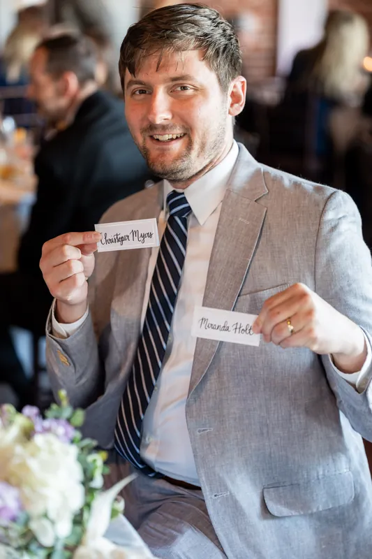 Tim holds up two place cards with guest names and grins widely at the reception, white flowers visible nearby.