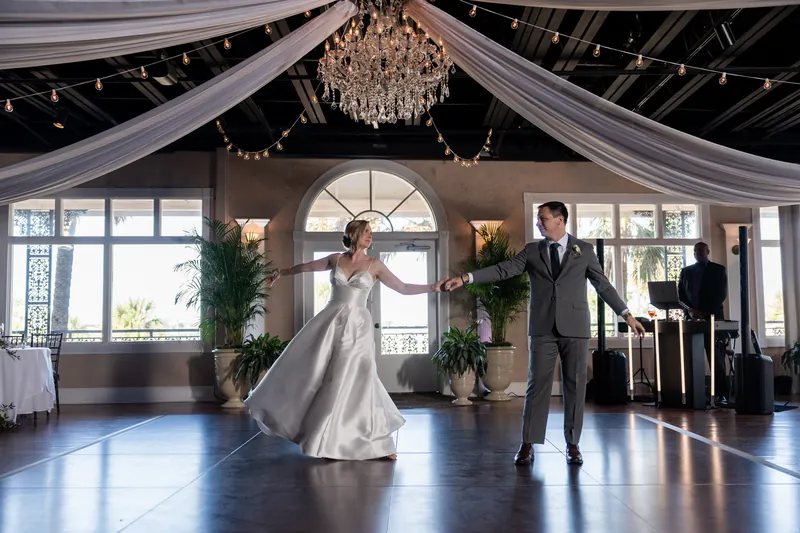 Sarah in a white gown and Tim in a gray suit perform their first dance under a draped ceiling with a crystal chandelier.
