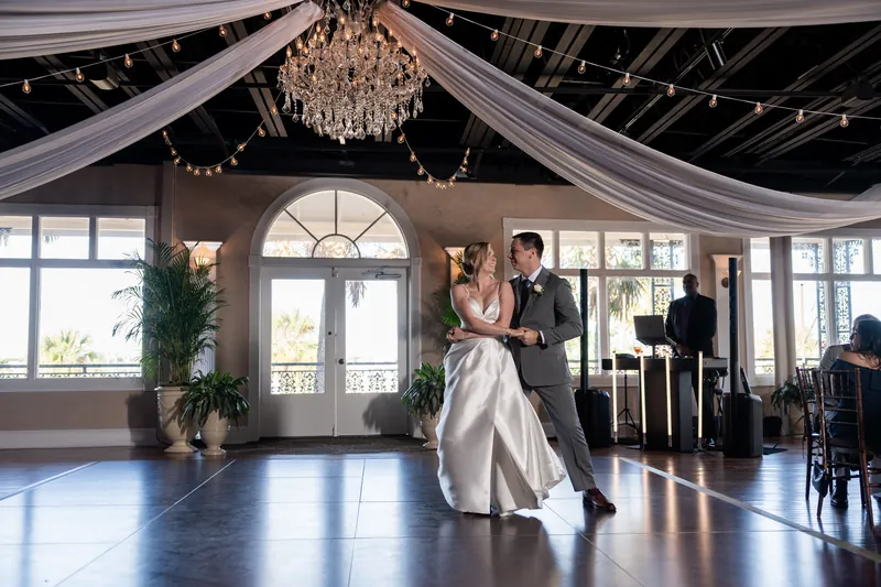 Sarah and Tim waltz across the polished dance floor beneath dramatic fabric draping and a crystal chandelier.