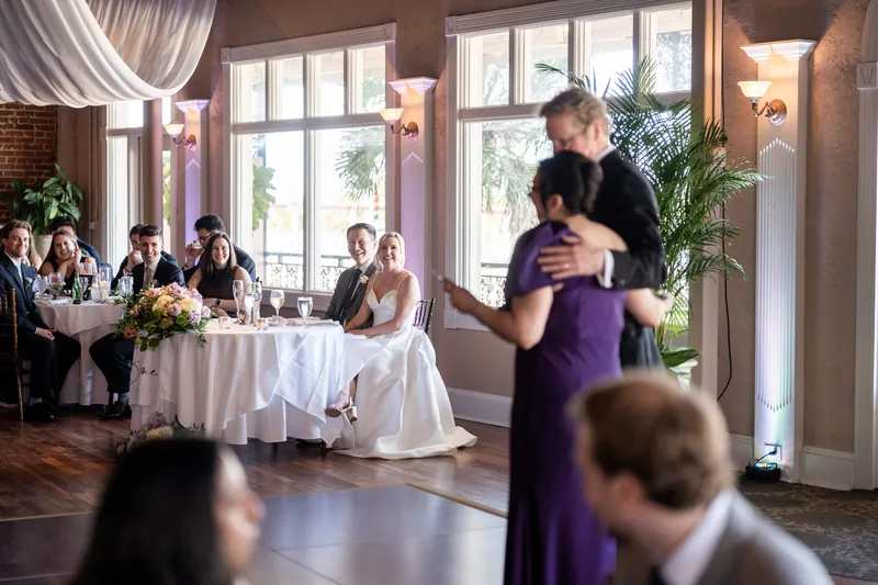 Tim and Sarah dance together while guests watch from tables in the bright, plant-lined reception hall.