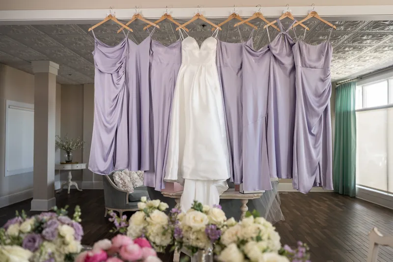 A white wedding dress hangs center among lavender bridesmaids' gowns on a rod, floral arrangements below.