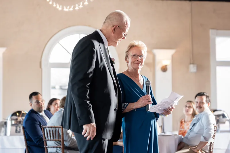 An older man and woman stand and speak while holding papers, addressing guests scattered throughout the reception space.