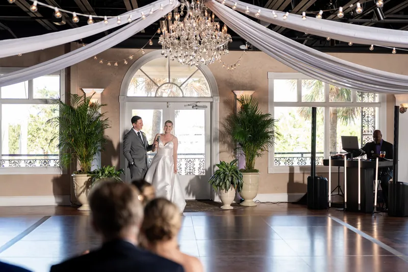 Sarah and Tim stand at the altar beneath a chandelier and draped fabric, with guests seated in the foreground at The White
