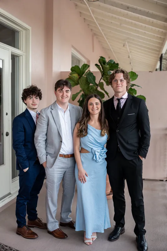 Four young people pose together indoors, with two in suits and one in a light blue dress.