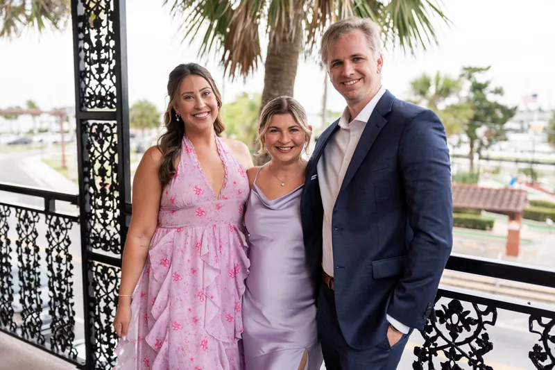 Three adults stand on a terrace with ornate black railings and a waterfront view behind them.