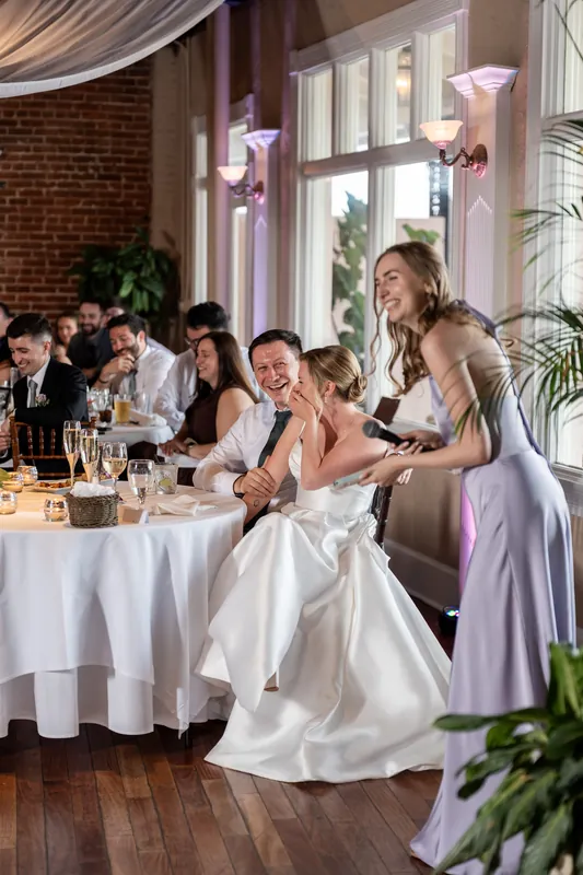 A woman in a white wedding dress sits at a reception table while another woman in lavender stands beside her speaking.