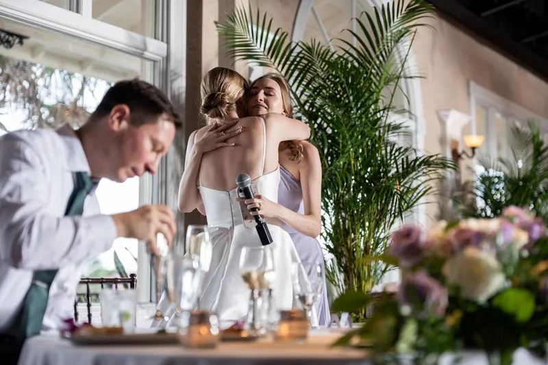 Tim leans toward Sarah at an indoor reception table decorated with greenery and white flowers.