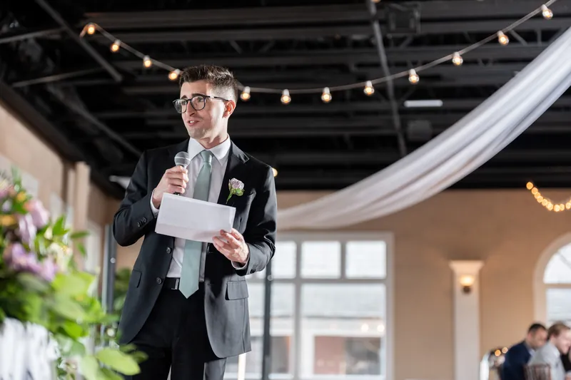 Tim holds a speech card and speaks into a microphone while standing in The White Room's soaring space, with string lights