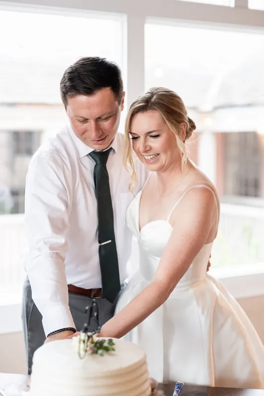 Sarah and Tim cut their white tiered cake together, both smiling as they guide the knife downward.