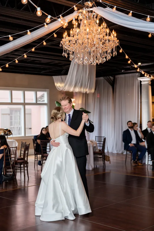 Sarah and Tim dance under a crystal chandelier strung between wooden beams, surrounded by seated guests.
