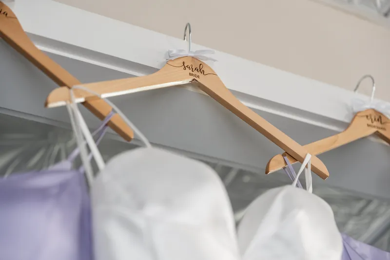 Wooden hangers with names engraved hang from a rod, with white and lavender garments draped beneath at The White Room.