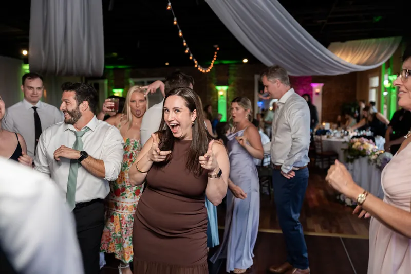 A guest in a brown maternity dress laughs while dancing among other reception attendees under draped fabric lighting.