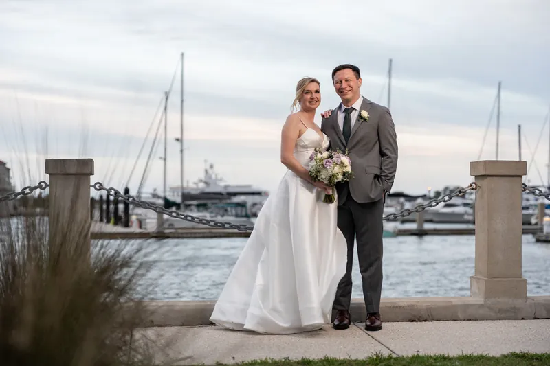Sarah and Tim stand together at the St. Augustine Bayfront with sailboats and water visible behind them, holding a white