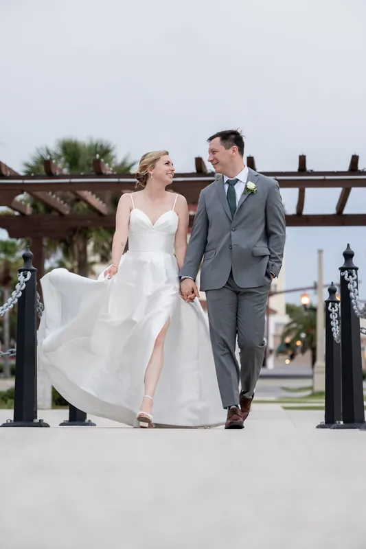 Sarah and Tim walk hand-in-hand along a wooden railing at the bayfront, Sarah's white gown flowing as they move.