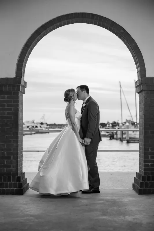 Sarah and Tim kiss framed by a brick archway with sailboats and water visible through the opening in black and white.