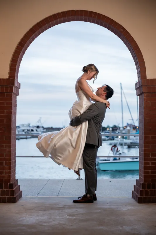 Tim lifts Sarah in his arms beneath a brick archway at the bayfront, with boats and water visible in the background.