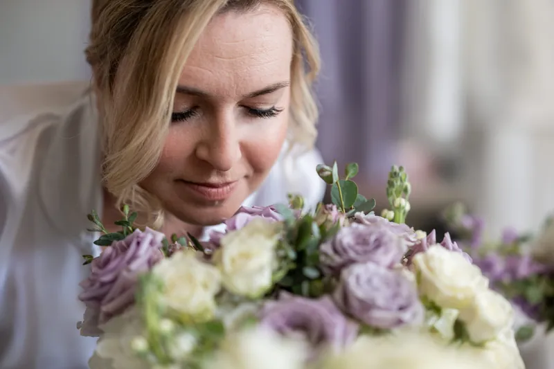 Sarah gazes down at a bridal bouquet of lavender and cream roses, smiling softly in getting-ready preparation.