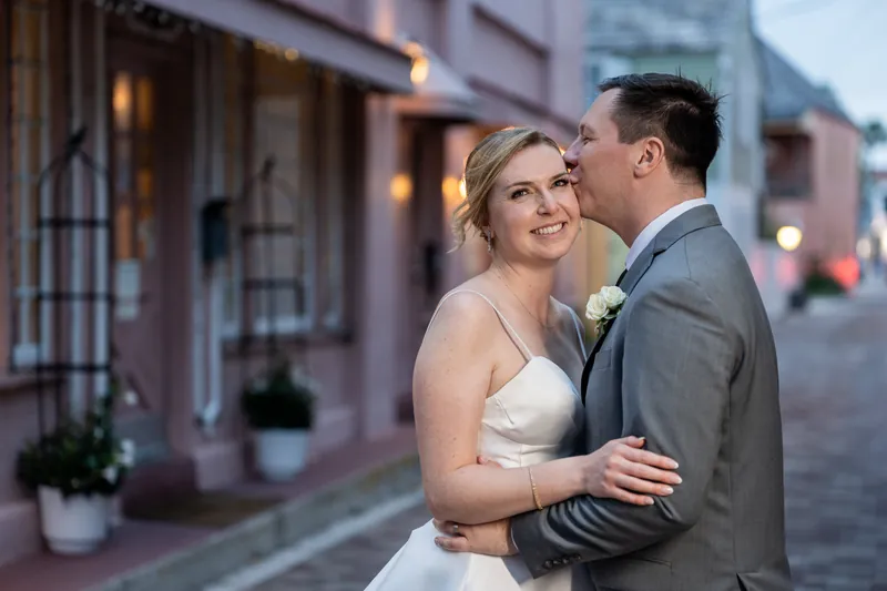 Tim embraces Sarah from behind on a cobblestone street at dusk, both smiling, with historic storefronts glowing behind them.