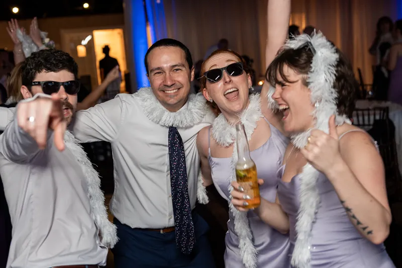 Four guests laugh together indoors wearing feather boas and sunglasses, holding drinks and celebrating at the reception.