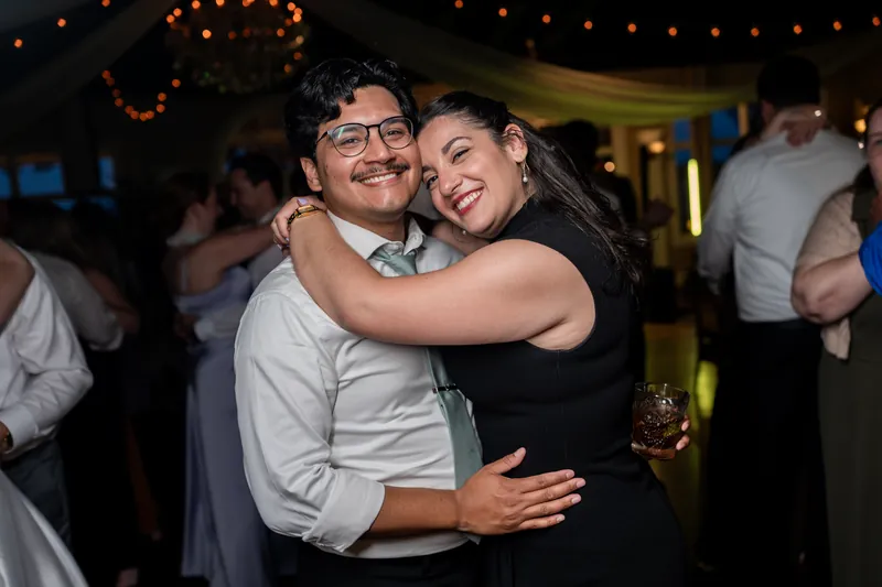 A man in glasses and light shirt hugs a woman in a black dress, both smiling during the reception.