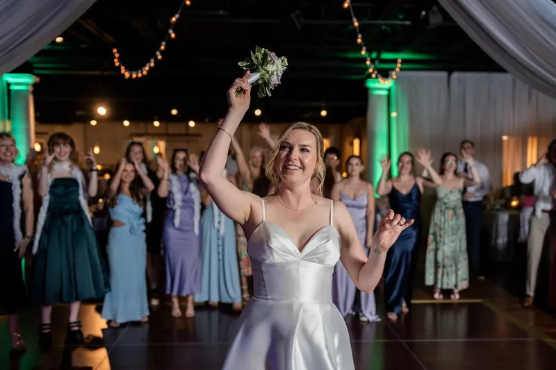 Sarah holds her bouquet high with one arm raised, beaming as guests surround her on the dance floor.
