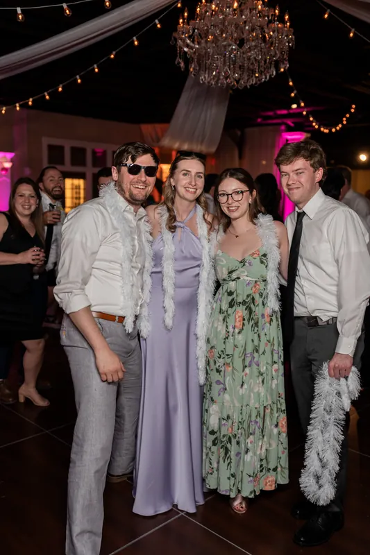 Four guests in mixed formal attire pose together at The White Room, one wearing a feather boa and sunglasses.