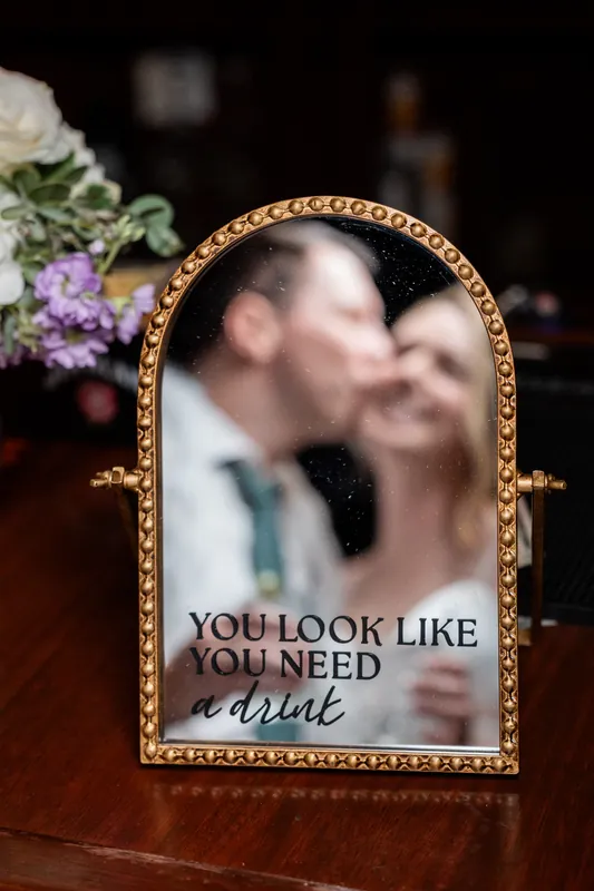 A gold-framed mirror with text reads "You look like you need a drink" beside purple flowers.