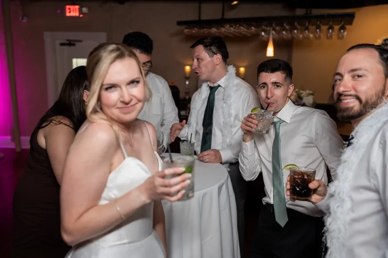 Sarah in a white strapless dress holds a cocktail while standing with male guests in the reception space.