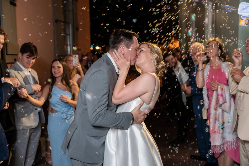 Sarah and Tim kiss while confetti falls around them, surrounded by smiling guests in formal attire at their reception.